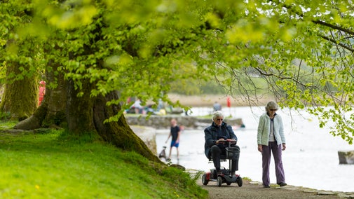 Person on mobility scooter and companion on a tree-lined lakeside path